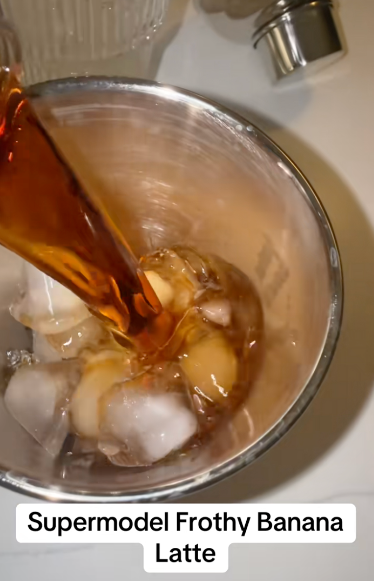 Pouring brown liquid over ice and banana slices in a metal cup, with the caption "Supermodel Frothy Banana Latte" below