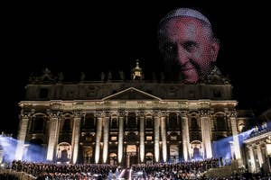 Nighttime event at St. Peter's Basilica with a light display of a large face in the sky, likely related to the event