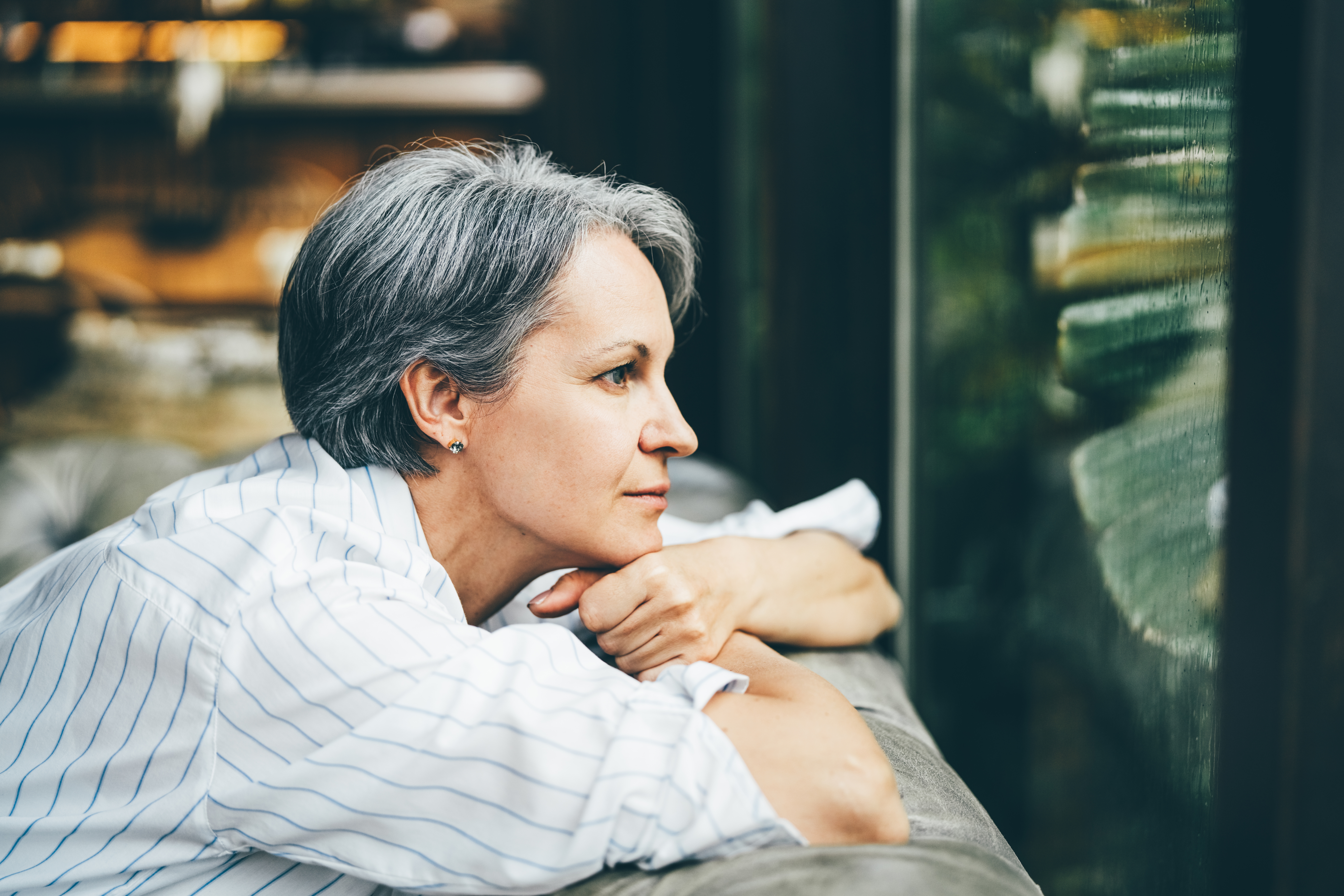 Person med kort hår i en stripet skjorte, hvilende hake på hendene, stirrer gjennomtenkt ut av vinduet