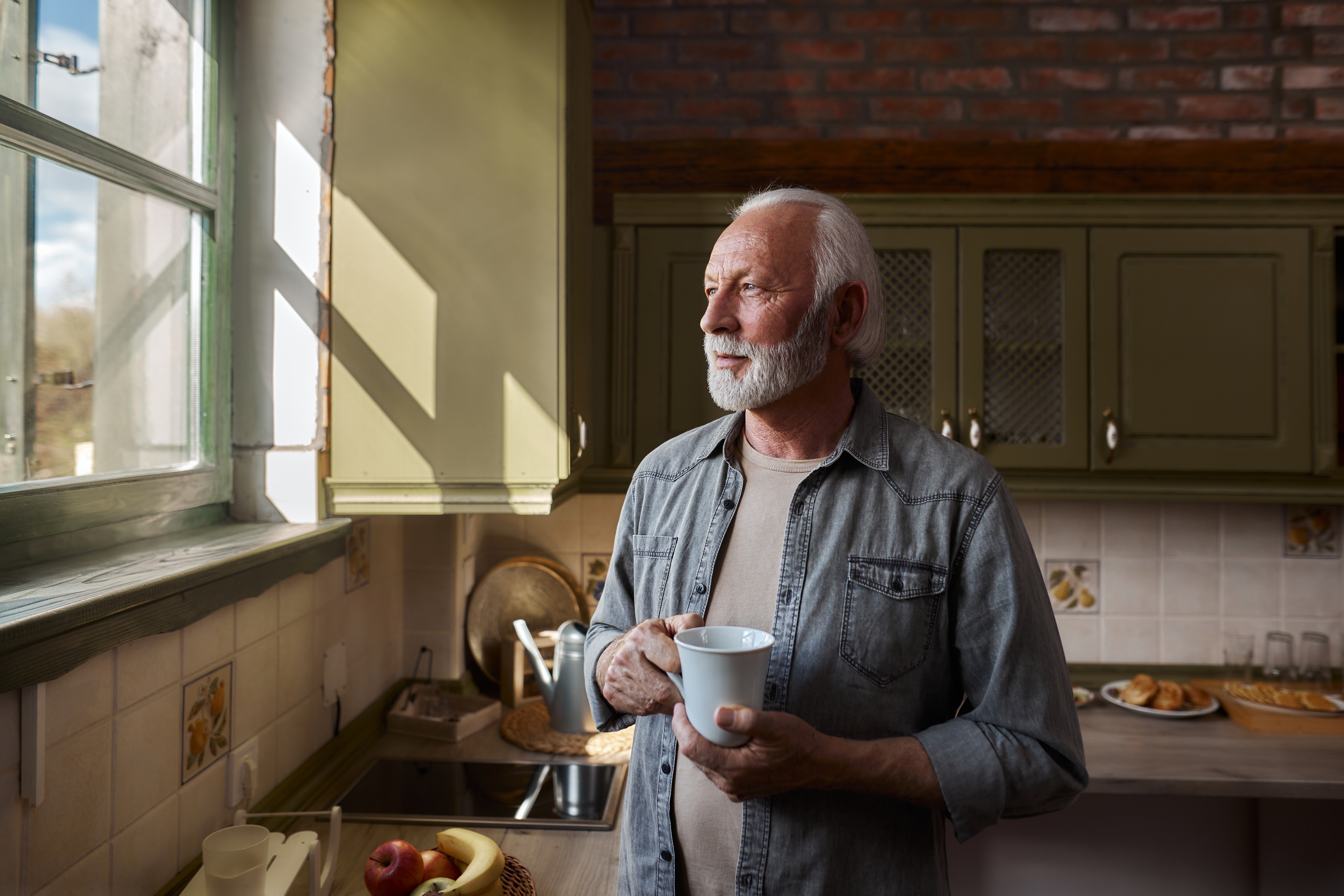 Eldre mann med skjegg holder et krus, og stirrer gjennomtenkt et kjøkkenvindu, omgitt av koselige hjemmekoselige elementer