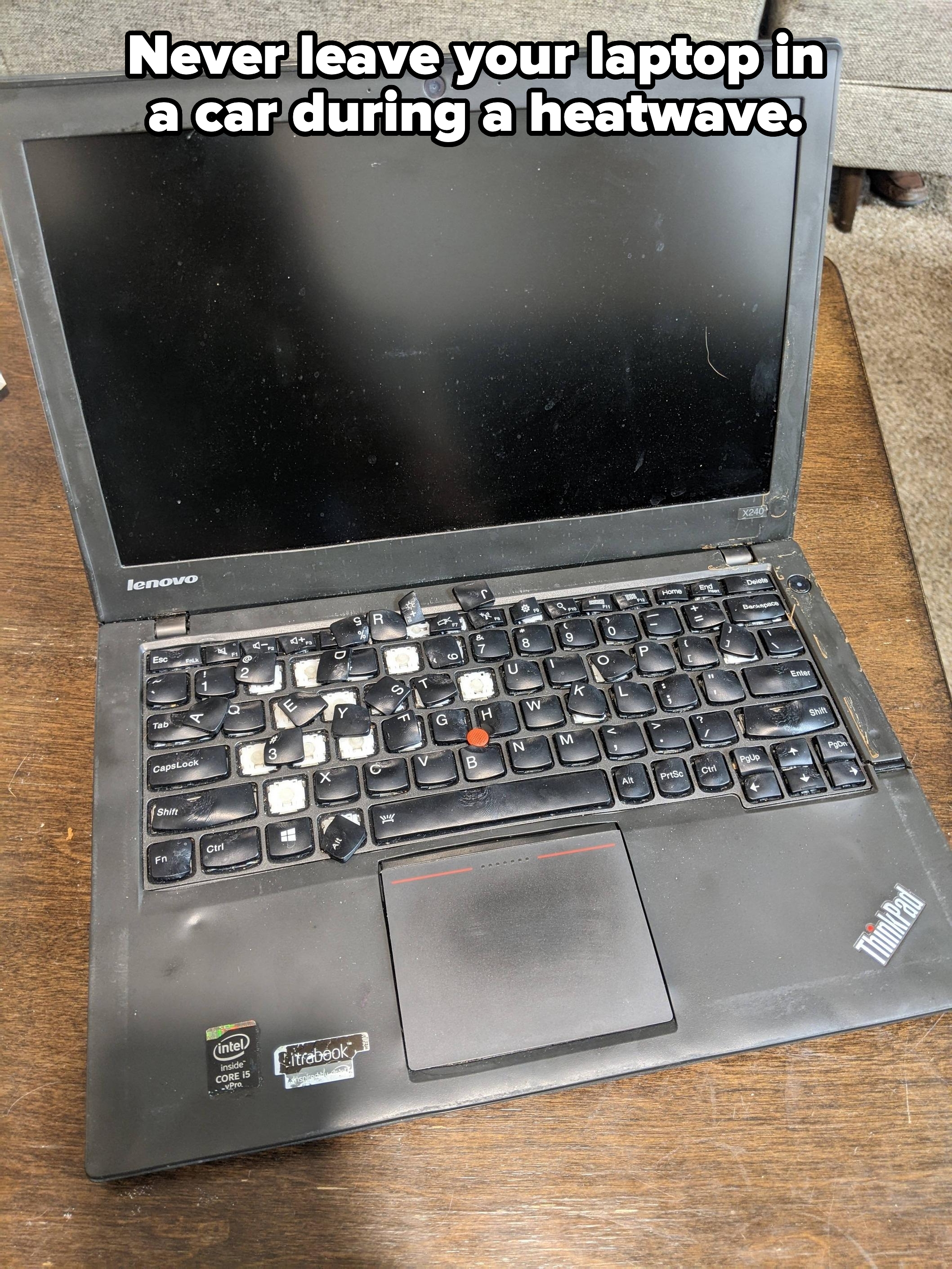 A well-used Lenovo laptop with a missing “J” key sits on a wooden table. The screen is off, and the keyboard shows signs of heavy use
