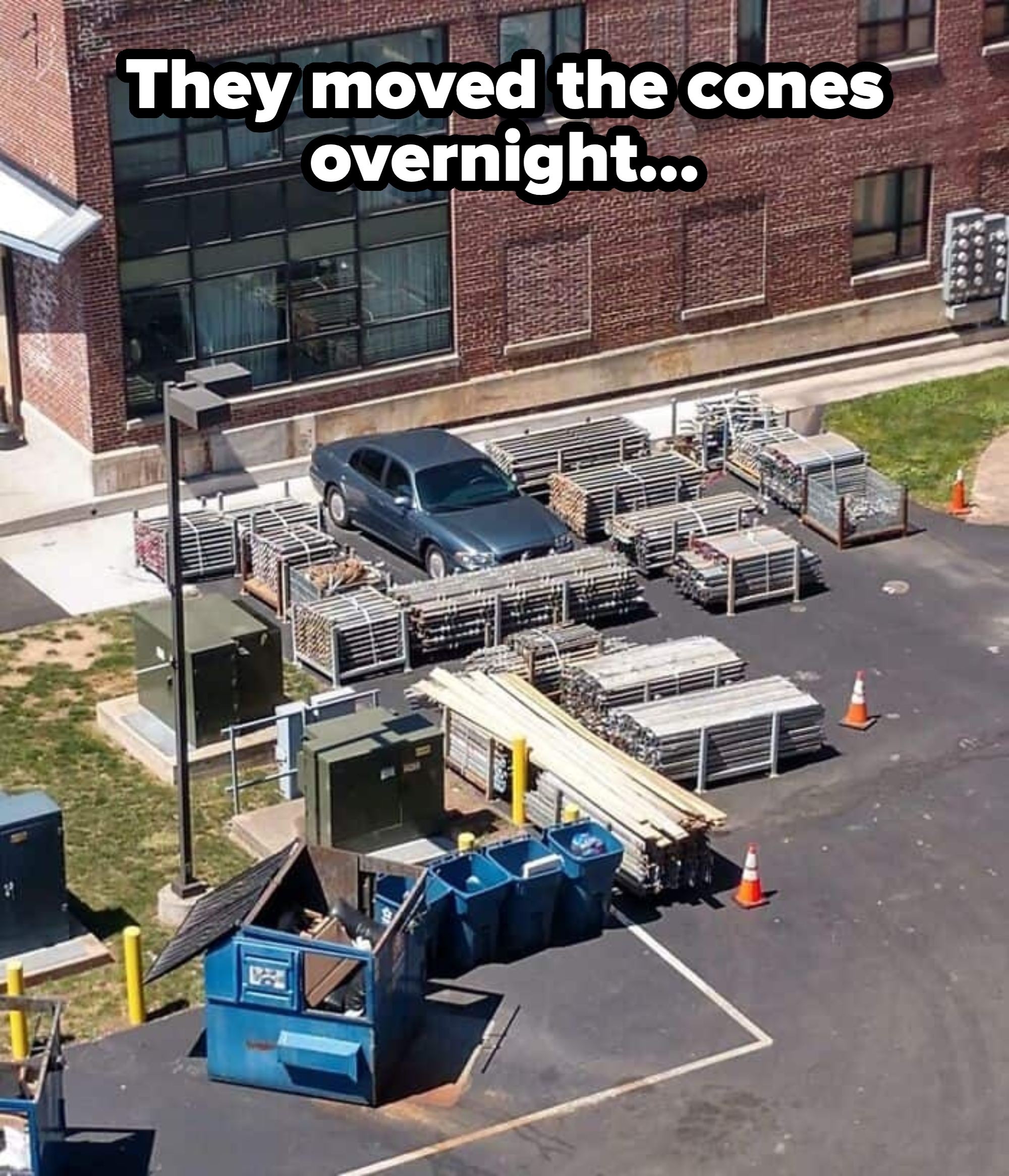 A car is parked in the middle of numerous stacked metal grids, surrounded by traffic cones and dumpsters in an urban area