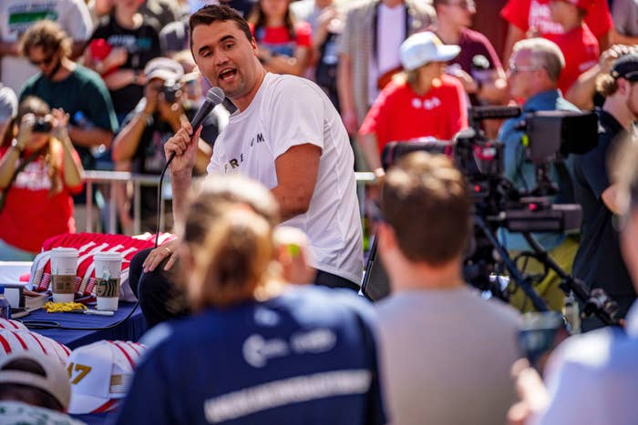 Charlie Kirk holds a microphone, speaking to a assemblage astatine an outdoor event. Camera unit and photographers are disposable successful the background