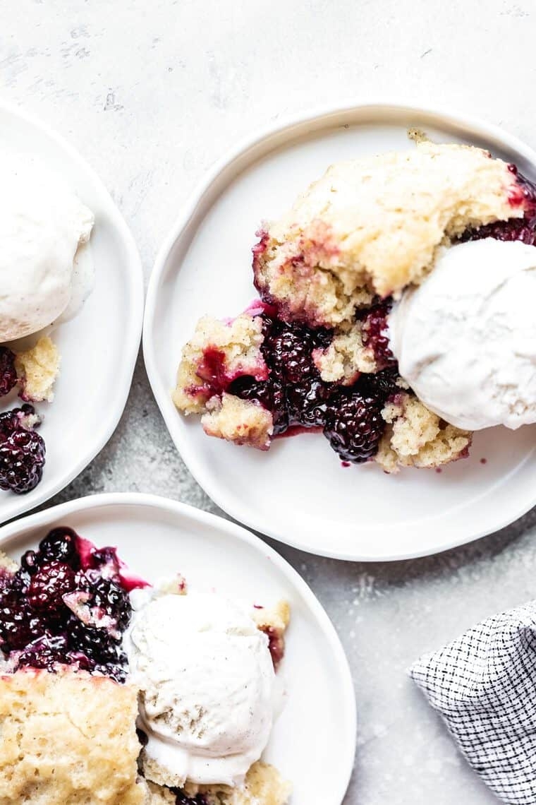Three plates of blackberry cobbler topped with scoops of vanilla ice cream. A partial view of a napkin is visible