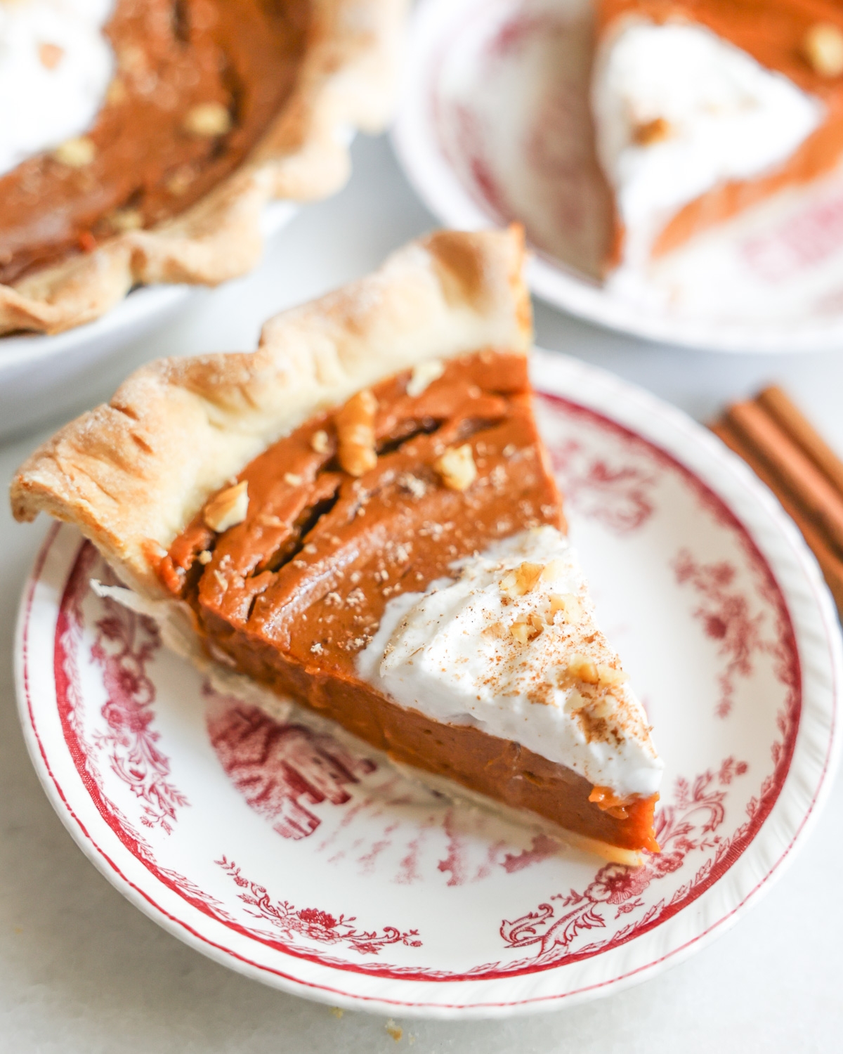 Slice of pumpkin pie with whipped cream on a decorative plate, next to two cinnamon sticks