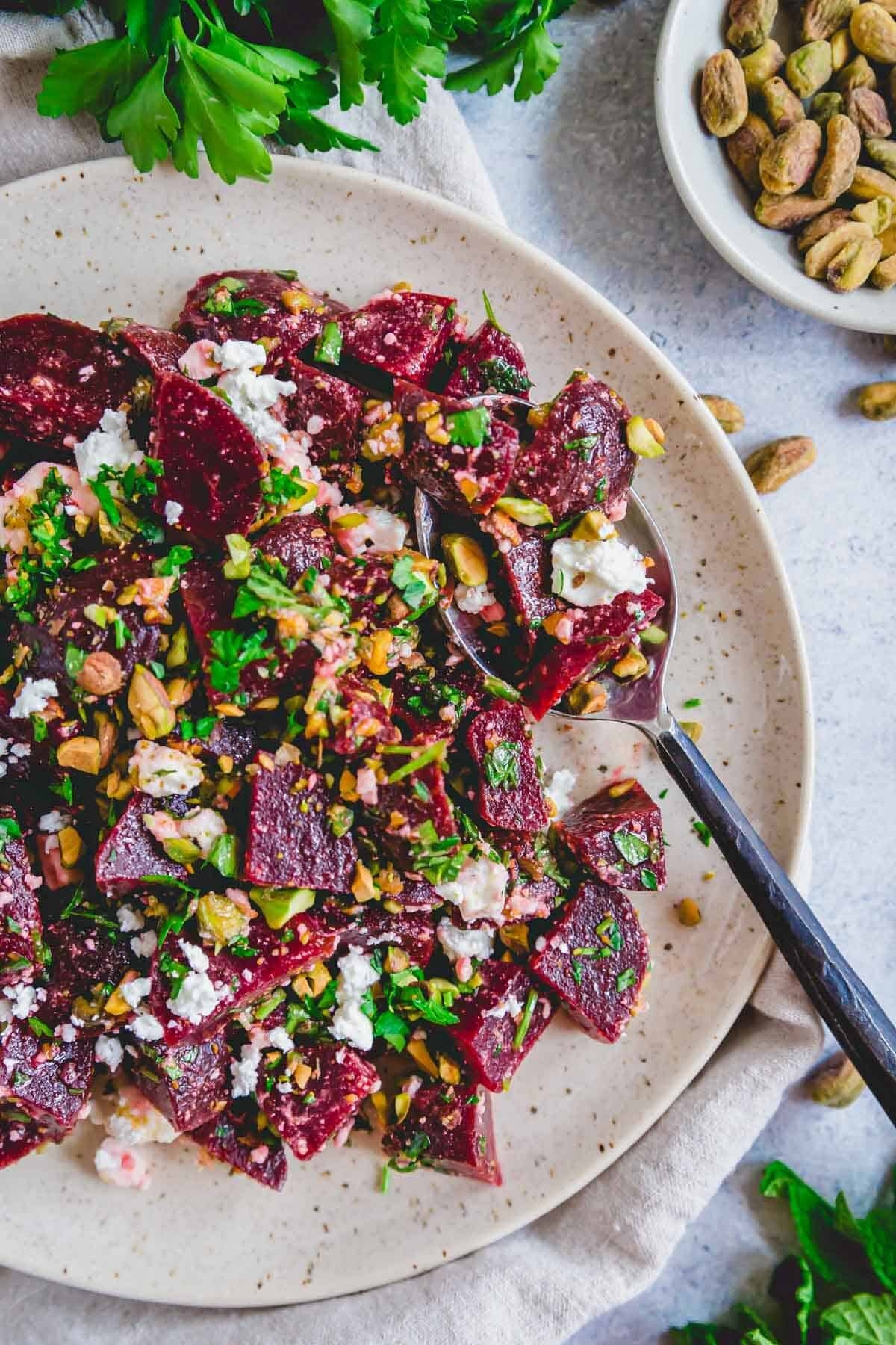 Plate of beet salad with feta, walnuts, and herbs, next to a bowl of pistachios. Fresh parsley garnishes the plate
