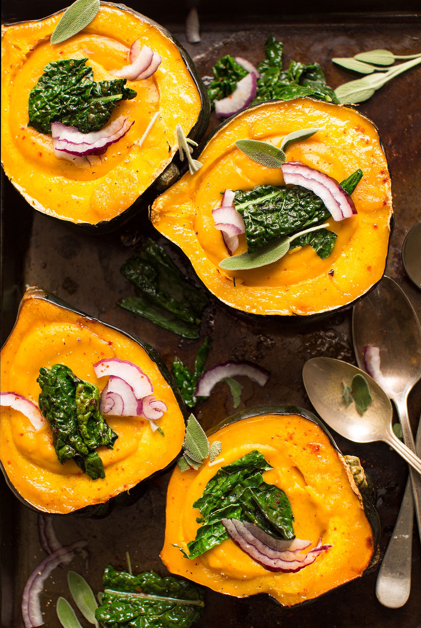 Four halved acorn squash filled with creamy soup, topped with kale and red onion slices, garnished with fresh sage leaves, and placed on a baking sheet