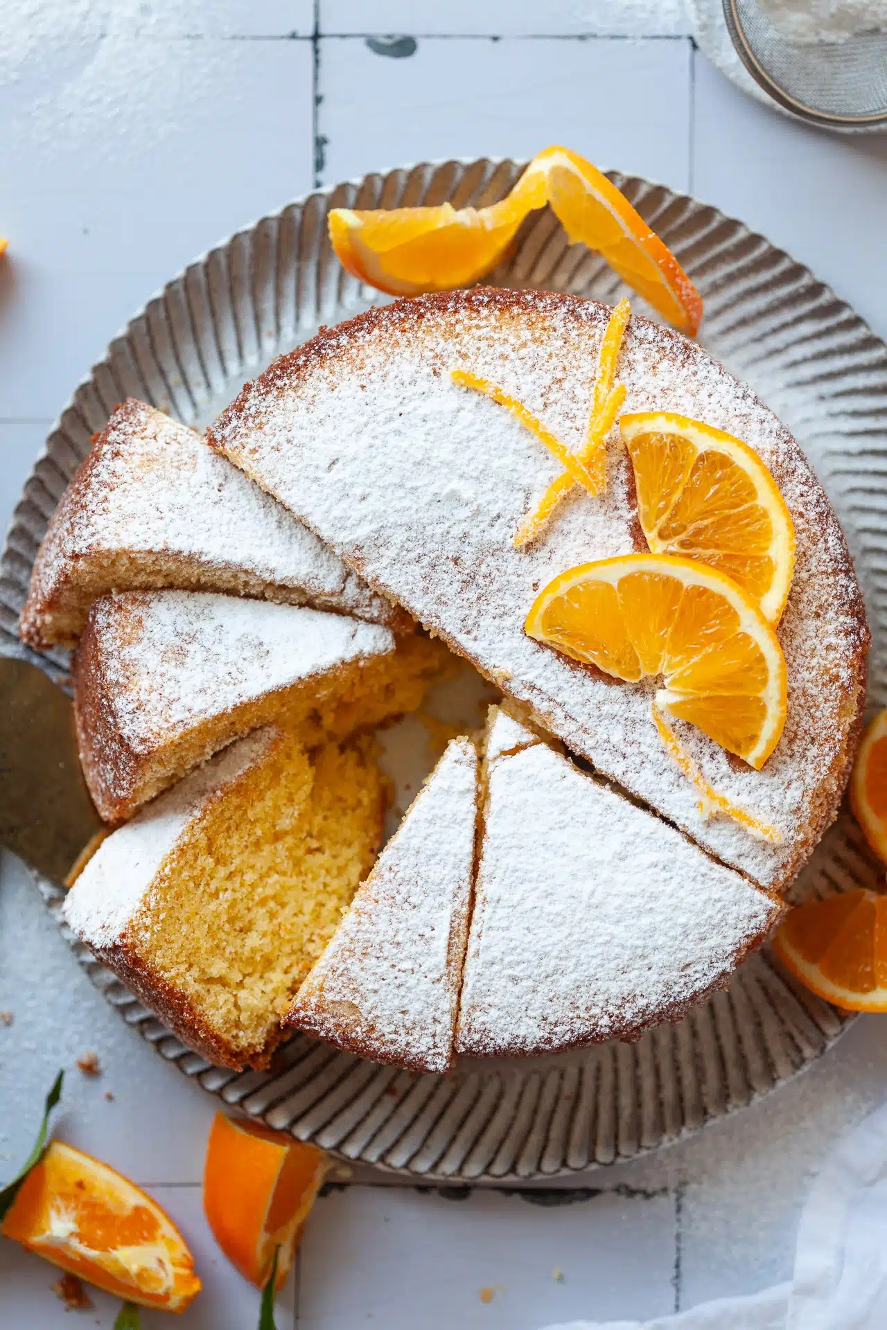 Cake topped with powdered sugar and orange slices, shown on a textured plate with a few slices cut and one partially removed