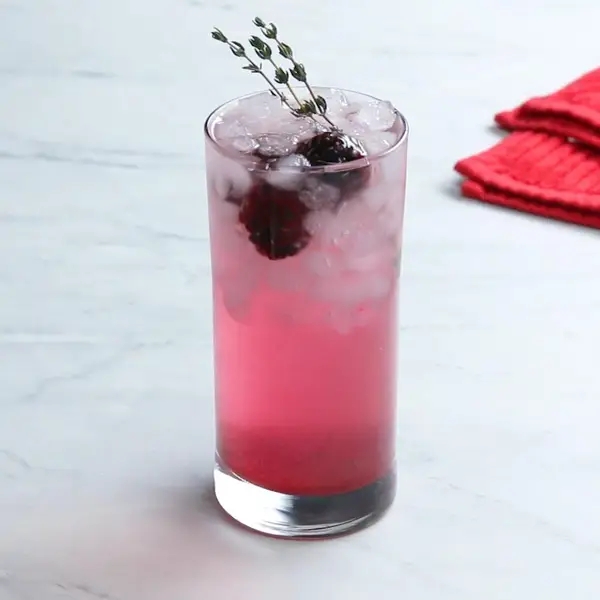 A pink cocktail in a tall glass with ice, garnished with a sprig of thyme and a blackberry, set on a marble surface next to a red napkin