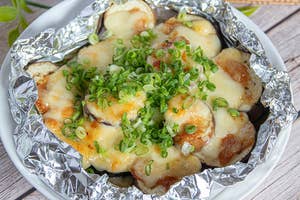 ホイルに包まれた焼きなすに、溶けたチーズと刻みネギがトッピングされている料理。