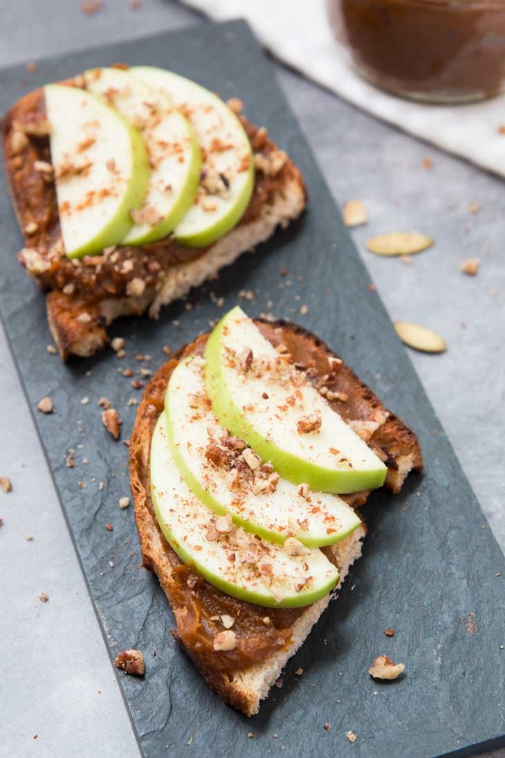 Two slices of toast topped with peanut butter, green apple slices, and crushed nuts on a slate board