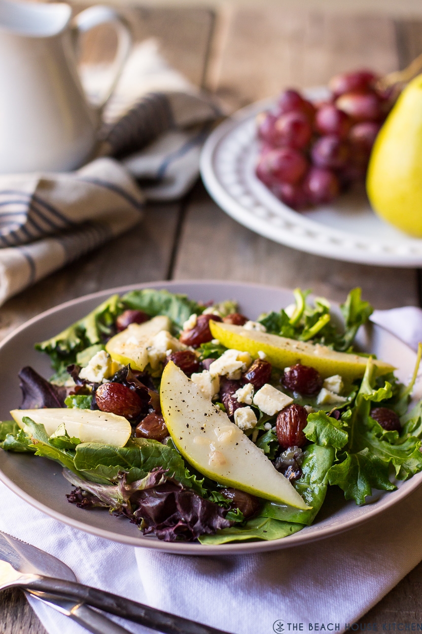 Salad on a plate with mixed greens, sliced pears, grapes, and crumbled cheese, on a wooden table with a napkin and fruit in the background