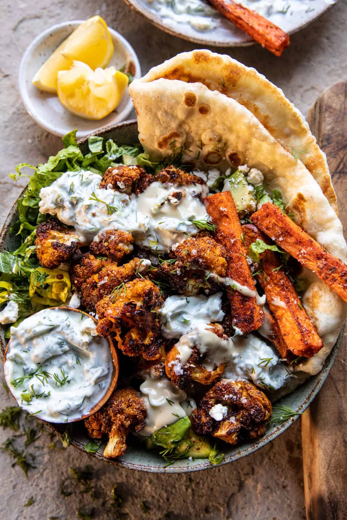 Bowl of roasted cauliflower and carrots drizzled with a creamy sauce, served with pita bread and lemon wedges