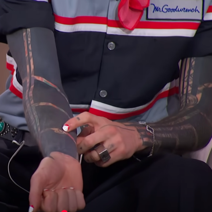 A person with tattooed arms and red manicured nails wearing a striped shirt with "Mr. Goodenough" embroidered, seated and pointing at their arm