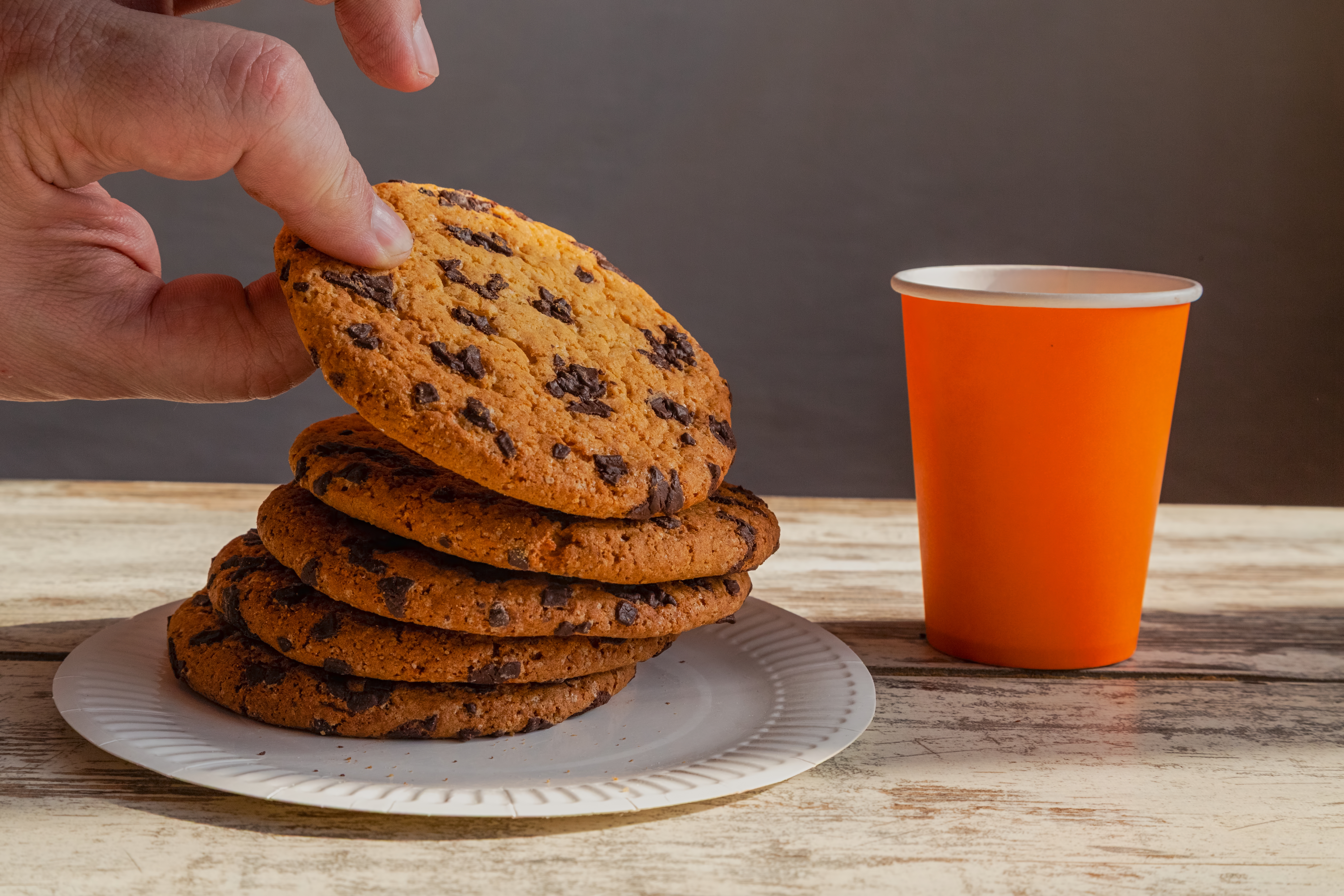 A hand picks up a chocolate chip cookie from a stack on a plate, next to an orange paper cup on a wooden table