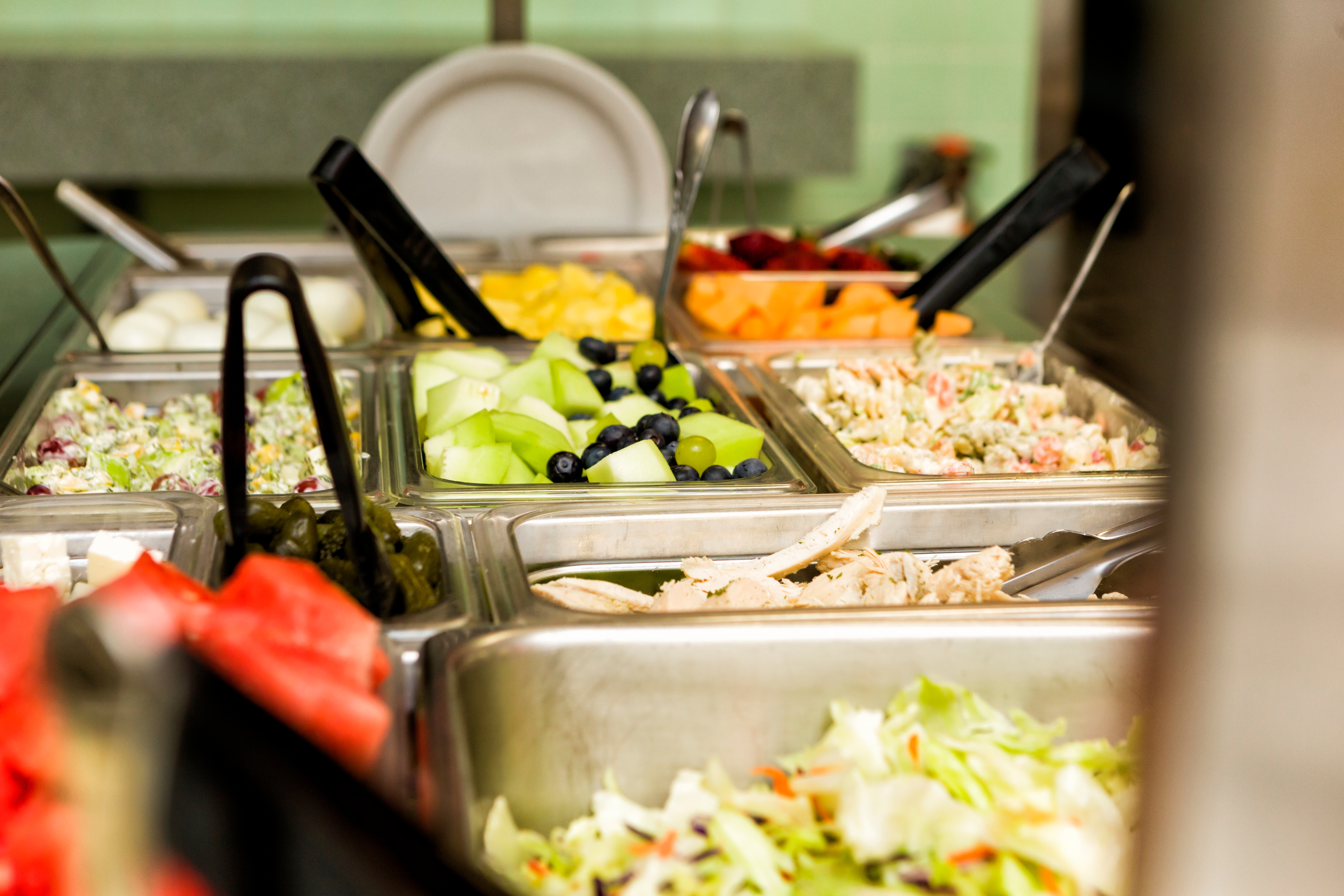 Salad bar with a variety of fresh ingredients including lettuce, melons, berries, and olives, displayed in metal trays with serving tongs
