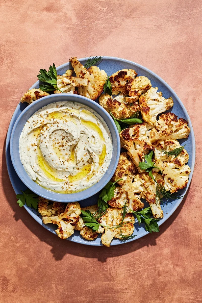 A plate of roasted cauliflower florets garnished with herbs, served with a bowl of creamy dip drizzled with olive oil