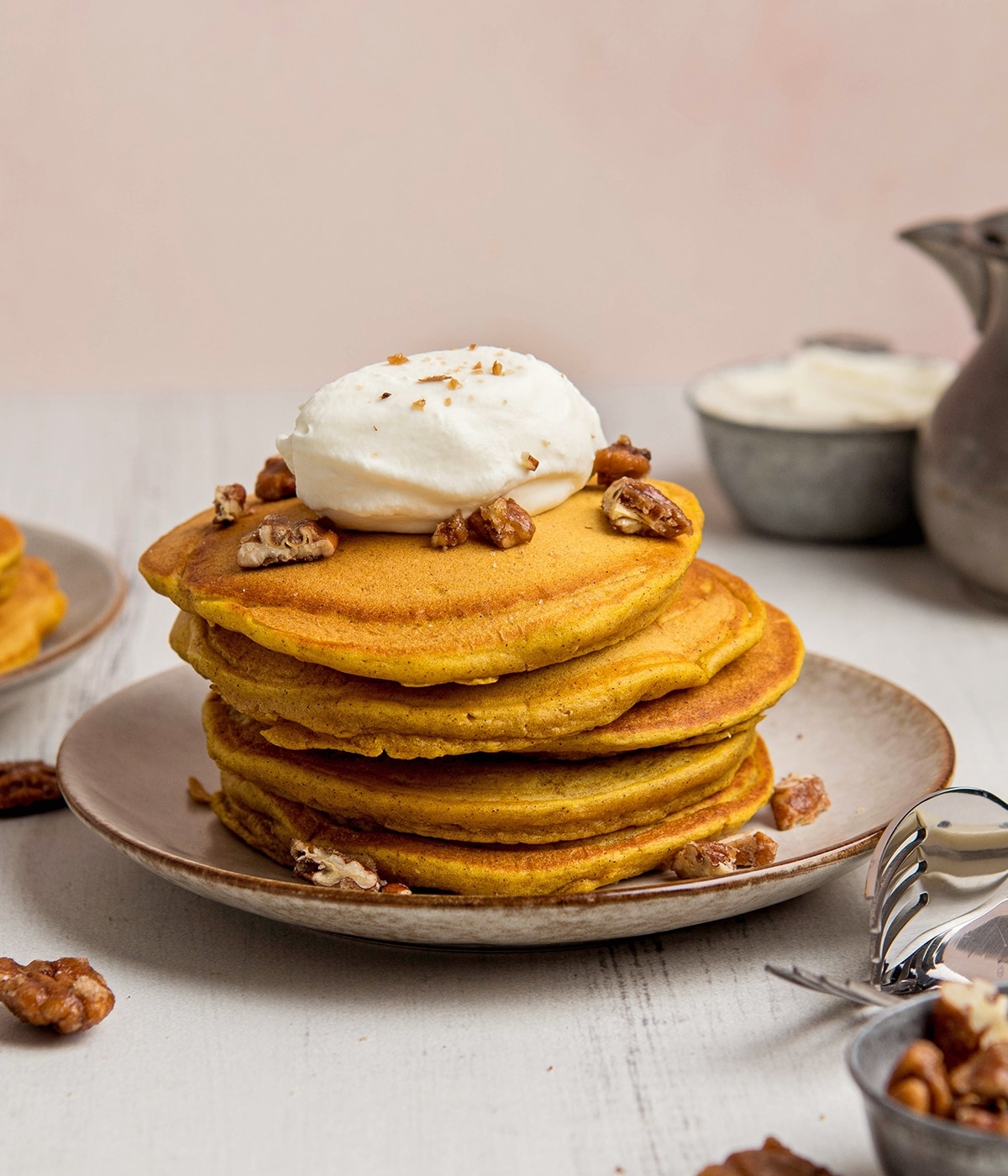Stack of pancakes topped with whipped cream and nuts on a plate, surrounded by a few utensils and a small dish