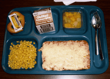 School lunch tray with pizza, corn, fruit cup, pretzel with mustard, chocolate milk, and utensils