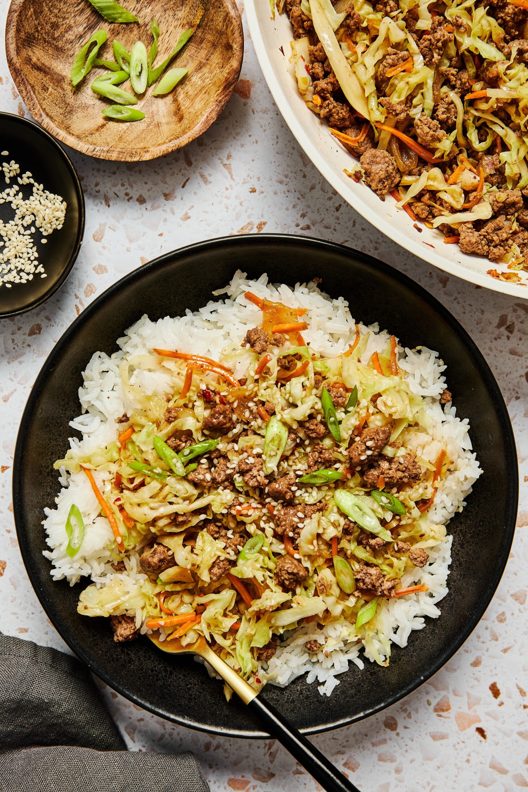 A bowl of rice topped with ground meat, shredded cabbage, carrots, green onions, and sesame seeds, with similar ingredients in the background