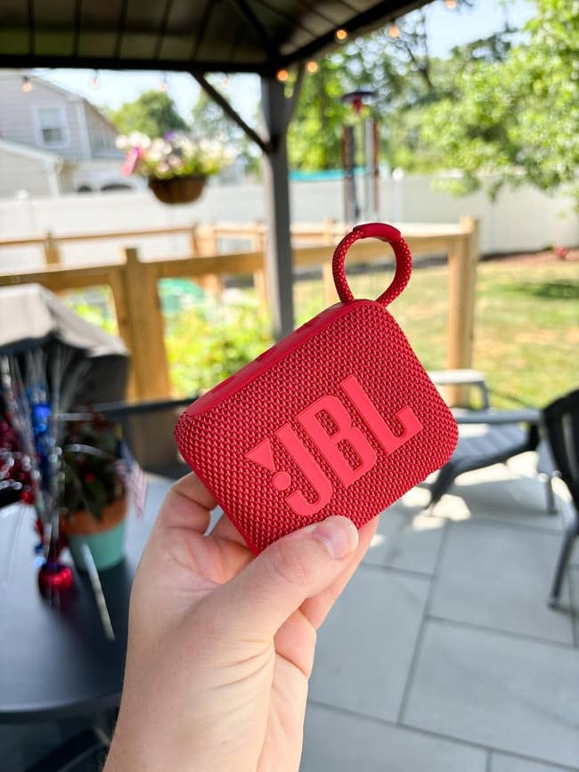 Person holding a small, textured JBL portable talker  with a loop handle, extracurricular  connected  a patio