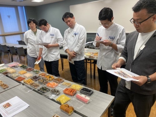 シェフたちが食品サンプルをテーブルに並べて評価している場面。