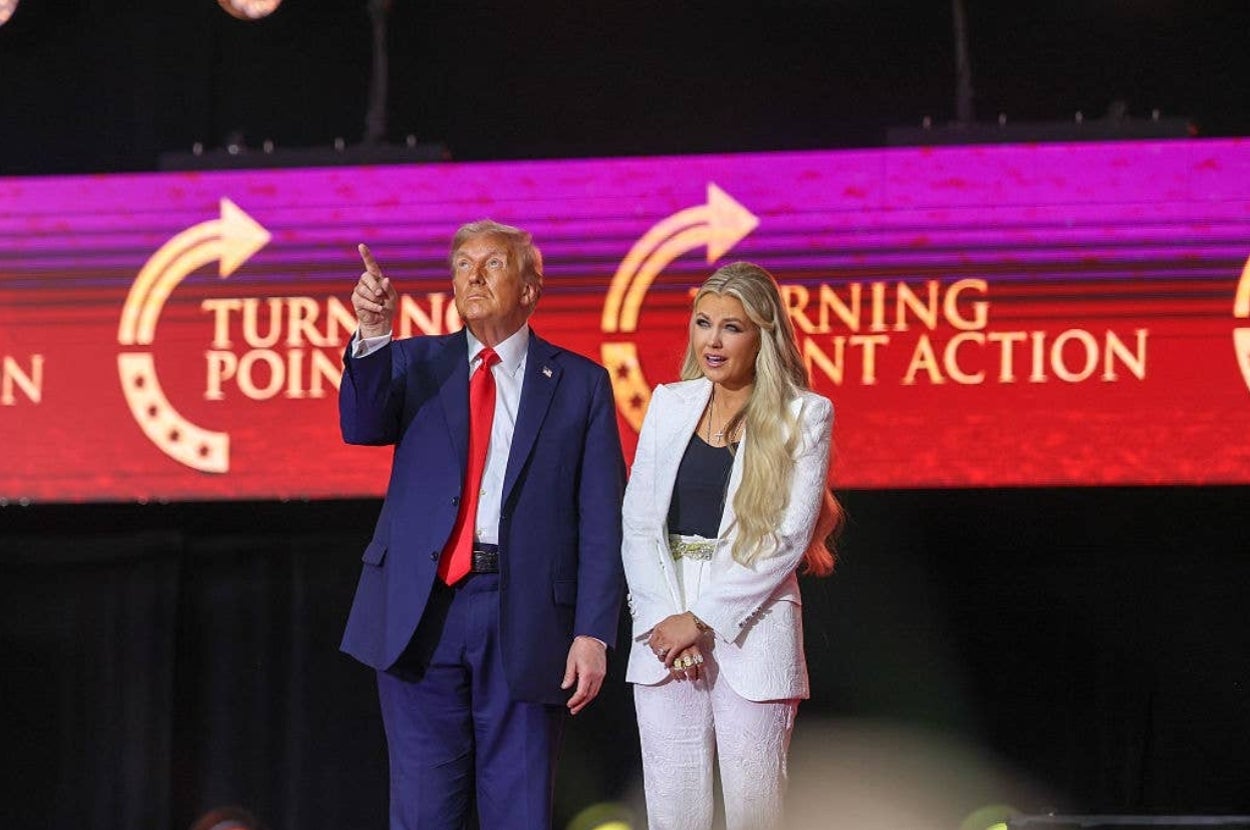Two people stand on stage at a Turning Point Action event. The man gestures forward while in a suit, and the woman is in a stylish pantsuit