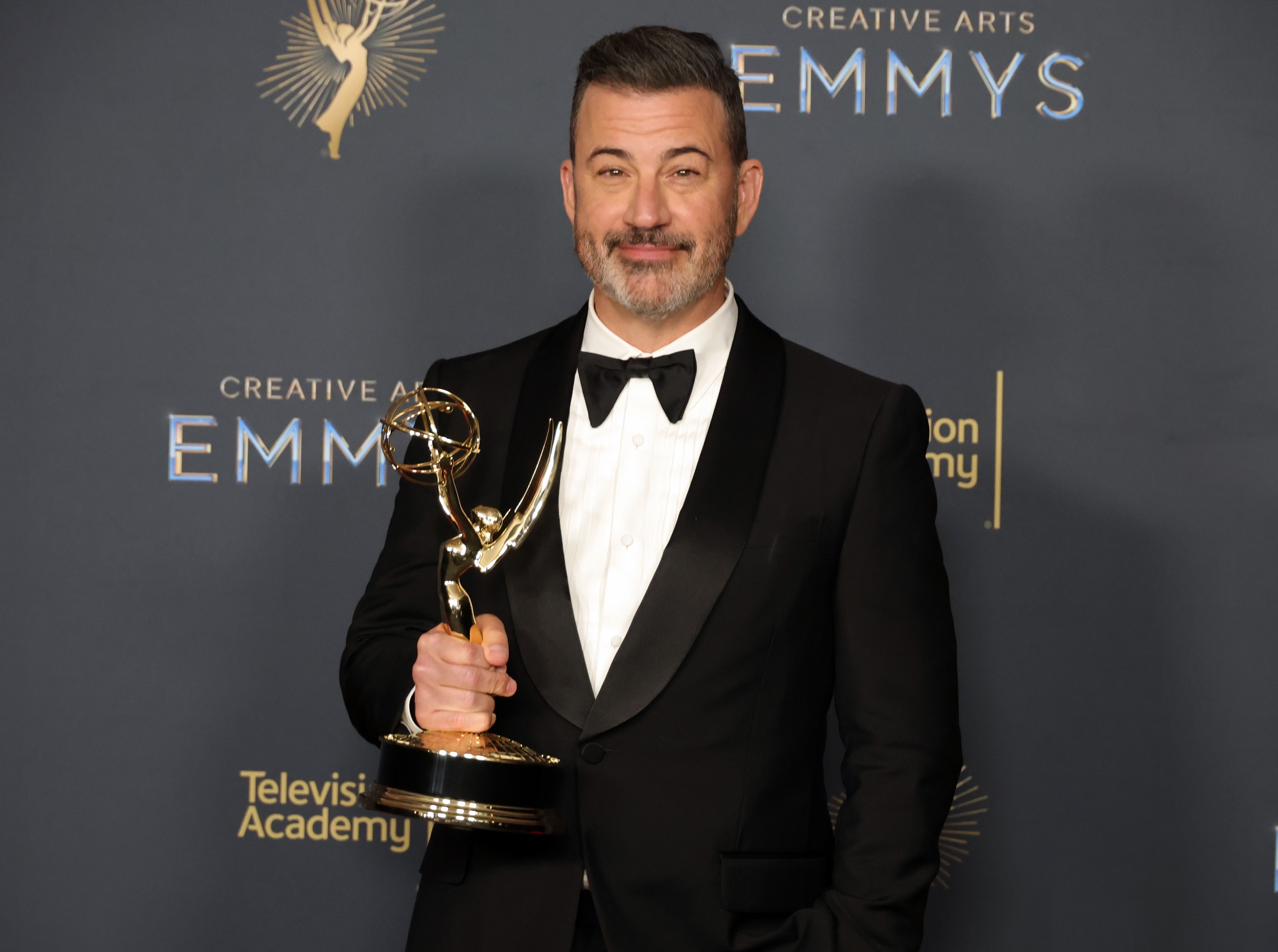 Jimmy Kimmel in a tuxedo holds an Emmy award while posing at a Creative Arts Emmys event
