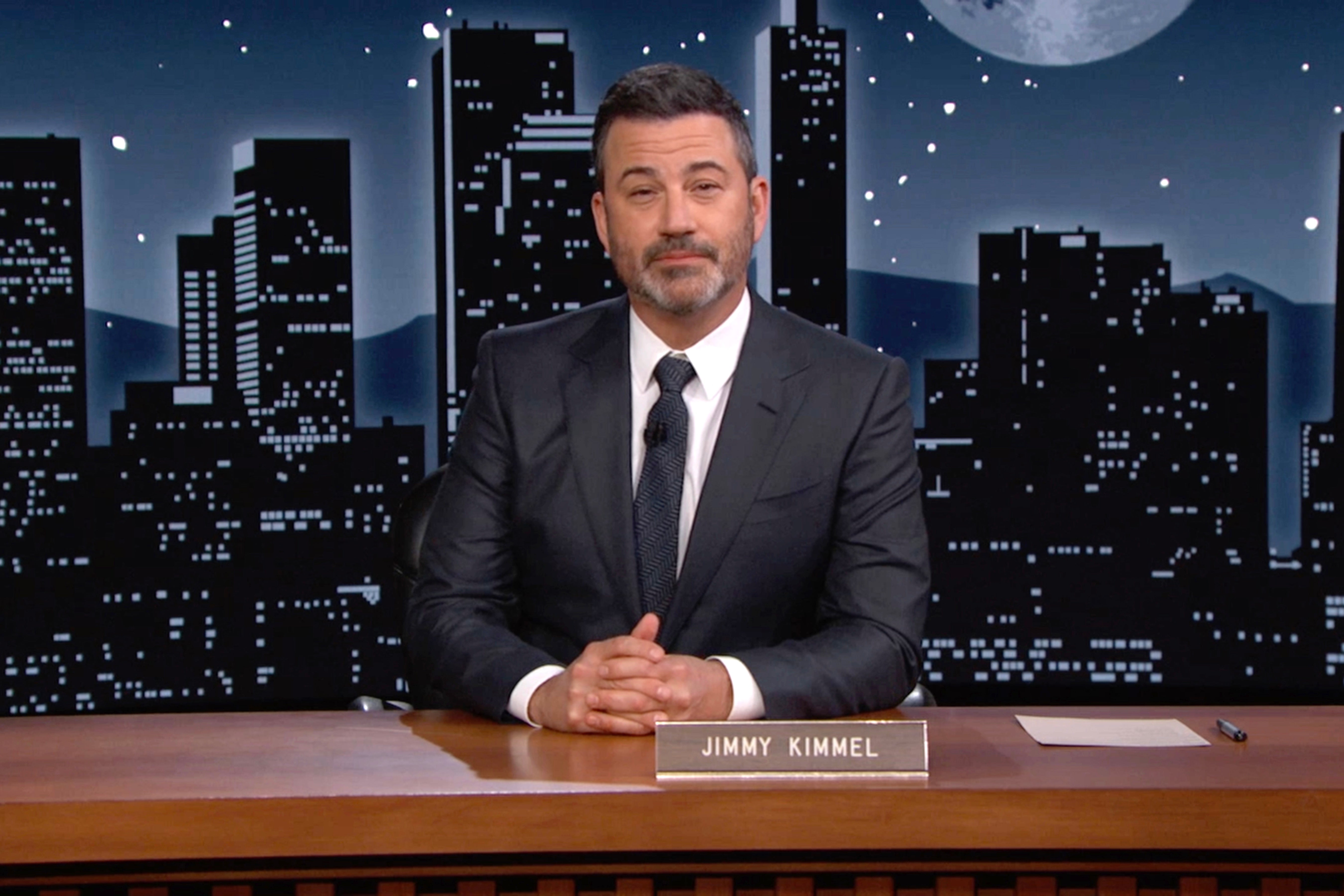 Talk show host in a suit sits at a desk with a city skyline backdrop and nameplate reading "Jimmy Kimmel."