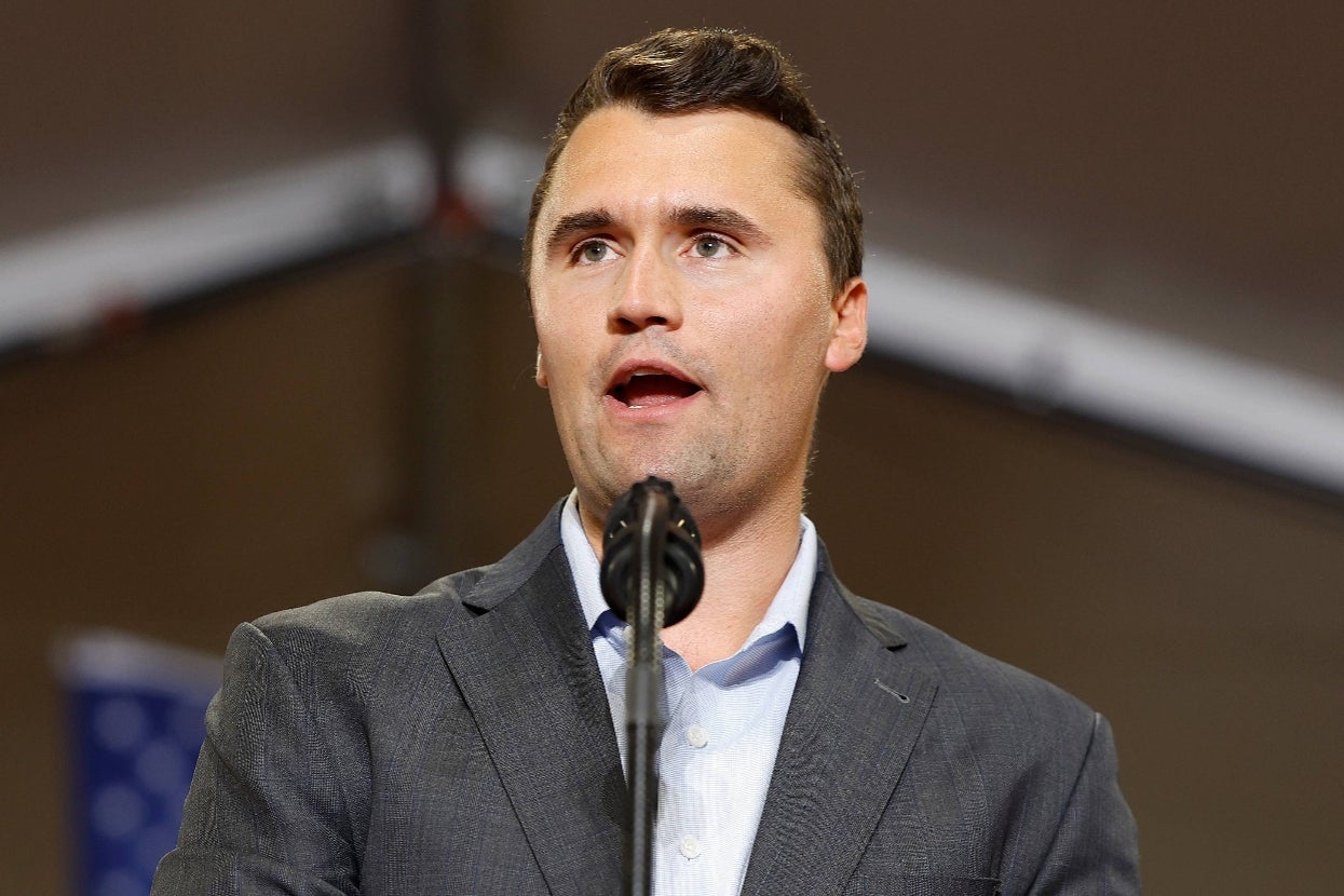 A man in a suit speaks at a podium with a microphone. A blurred flag is visible in the background