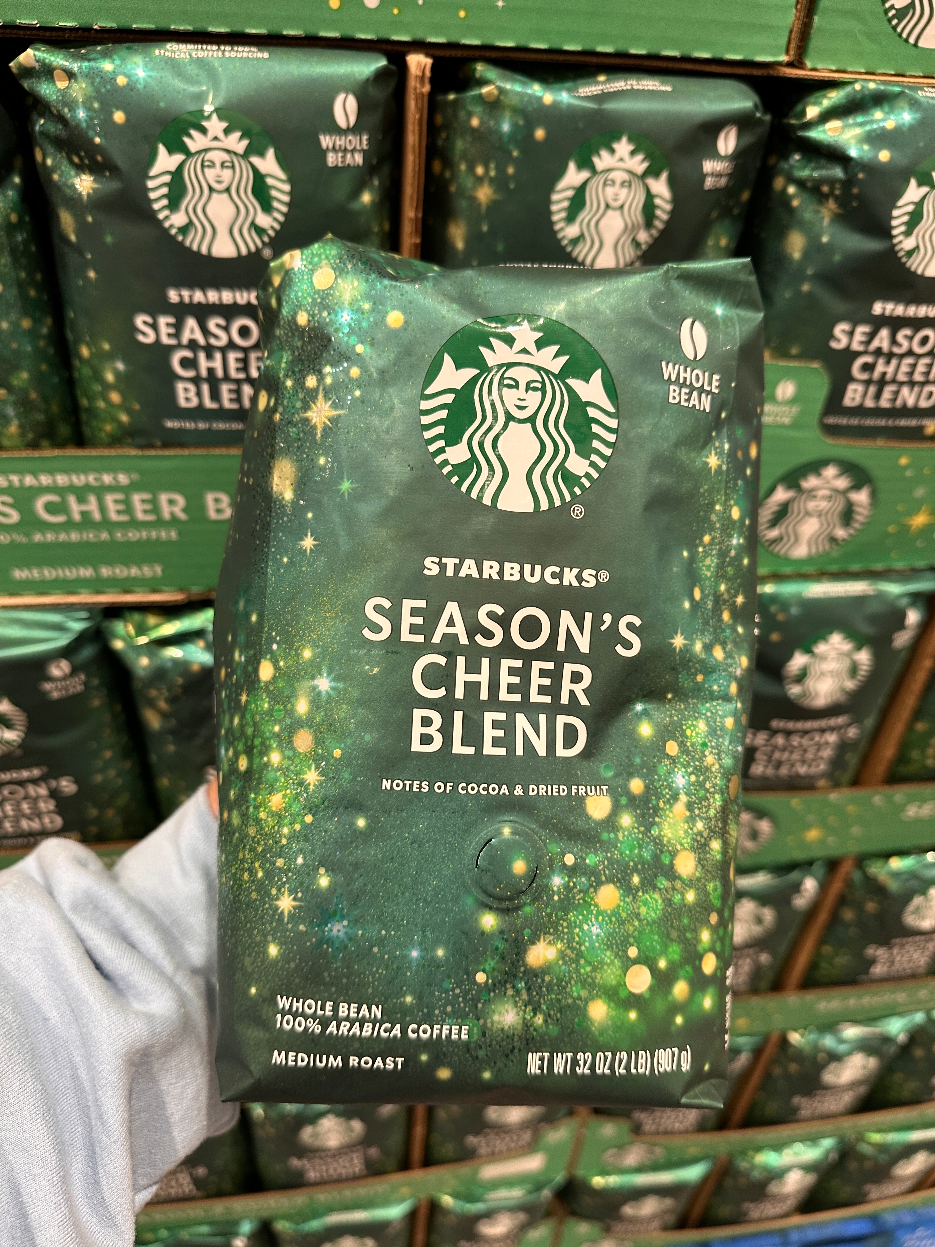 A hand holds a bag of Starbucks Season's Cheer Blend coffee, noting cocoa and dried fruit flavors. Other bags are stacked behind it