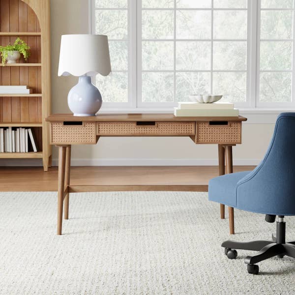 Modern wooden desk with cane accents, a blue lamp, bowls, and a blue chair on a light rug by large windows in a bright, home office setting