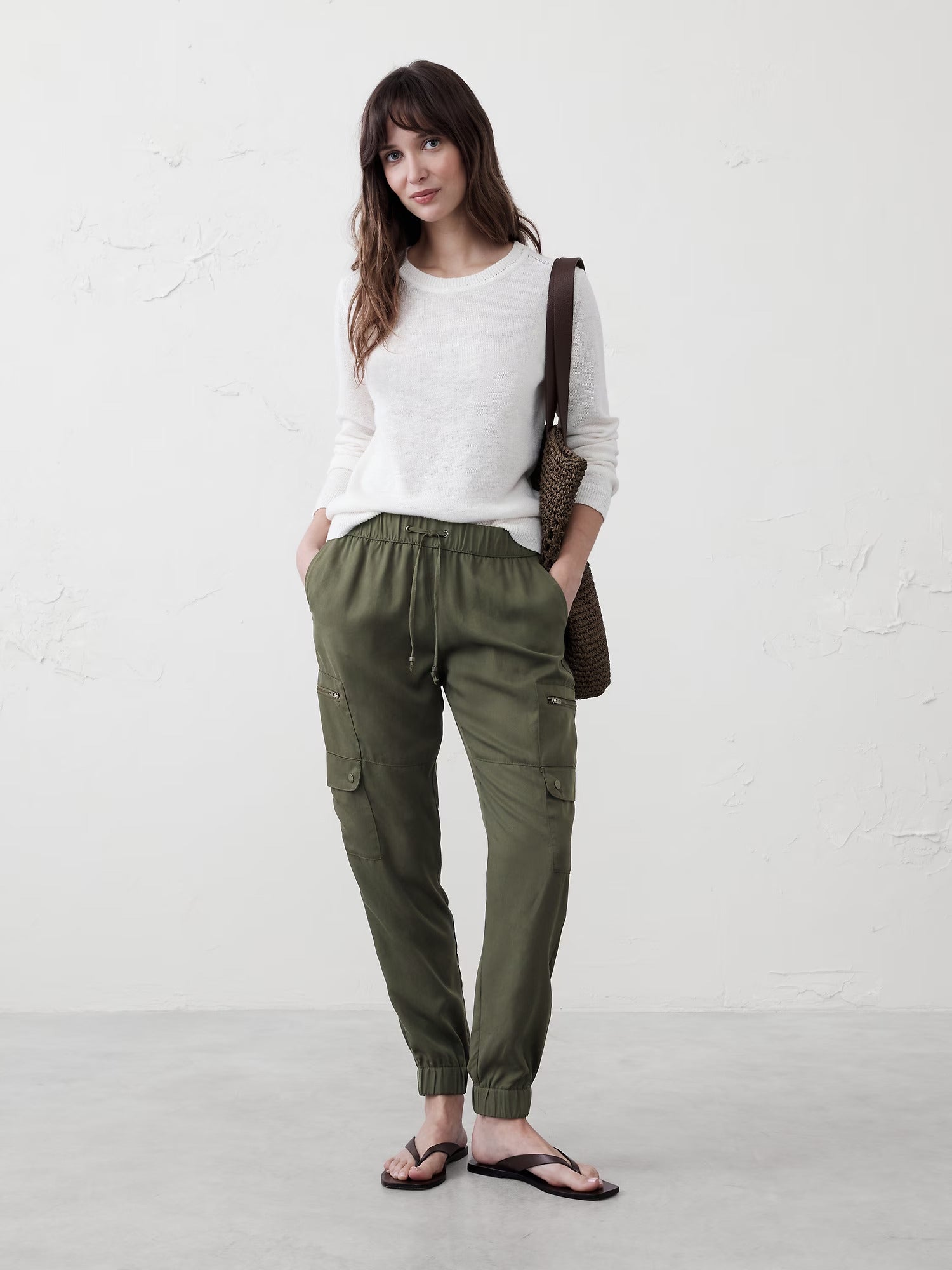 Person wearing a casual achromatic  sweater, greenish  cargo pants, and flip-flops, holding a woven bag