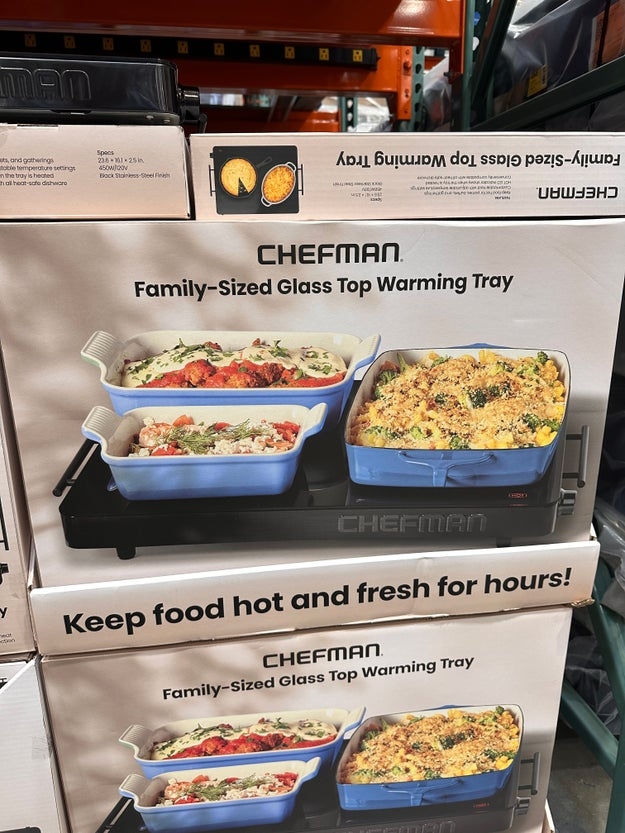 Boxes of Chefman family-sized glass top warming trays stacked in a store aisle. Packaging shows three trays with different dishes inside