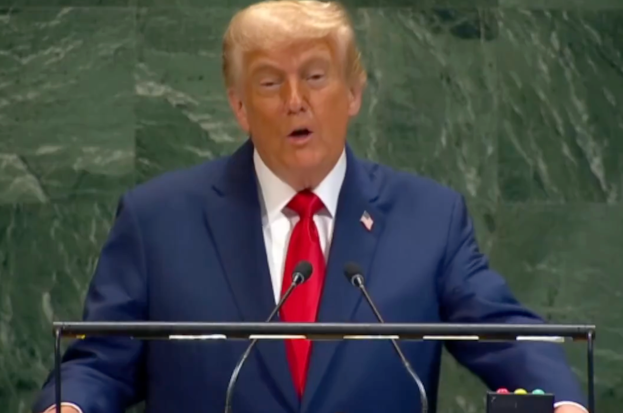 A person in a suit and red tie speaks at a podium with multiple microphones, looking engaged while addressing an audience