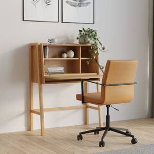 Wooden desk with shelving, decorated with a plant, mug, and books. A cushioned office chair is positioned in front of the desk. Minimalist design