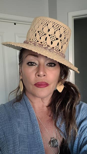 A reviewer wearing a woven chapeau with elaborate cutouts, connection earrings, a bluish textured top, and layered necklaces, posing indoors