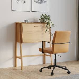 Compact wooden desk with fold-down panel, paired with a cushioned office chair on wheels, placed in a minimalistic room setting