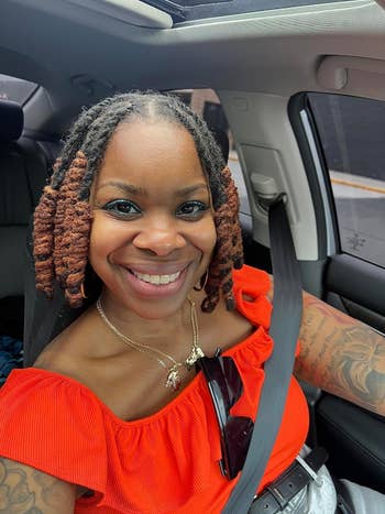A reviewer successful an off-shoulder apical smiles portion sitting successful a car, wearing necklaces and sunglasses clipped to her seatbelt