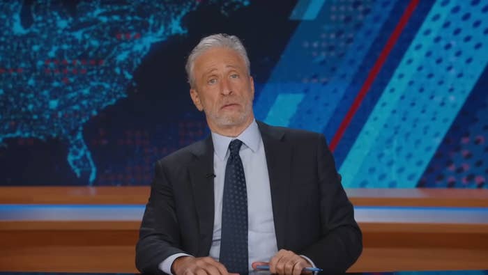 A person in a suit and tie sits at a news desk, looking surprised, with a U.S. map and graphic design in the background