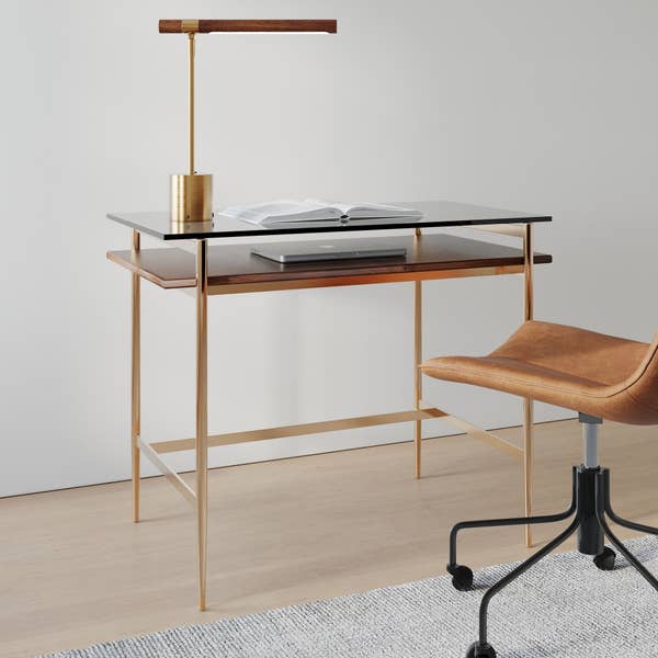 Sleek, modern writing desk with a glass surface, paired with a brown leather office chair and a stylish gold and wood desk lamp