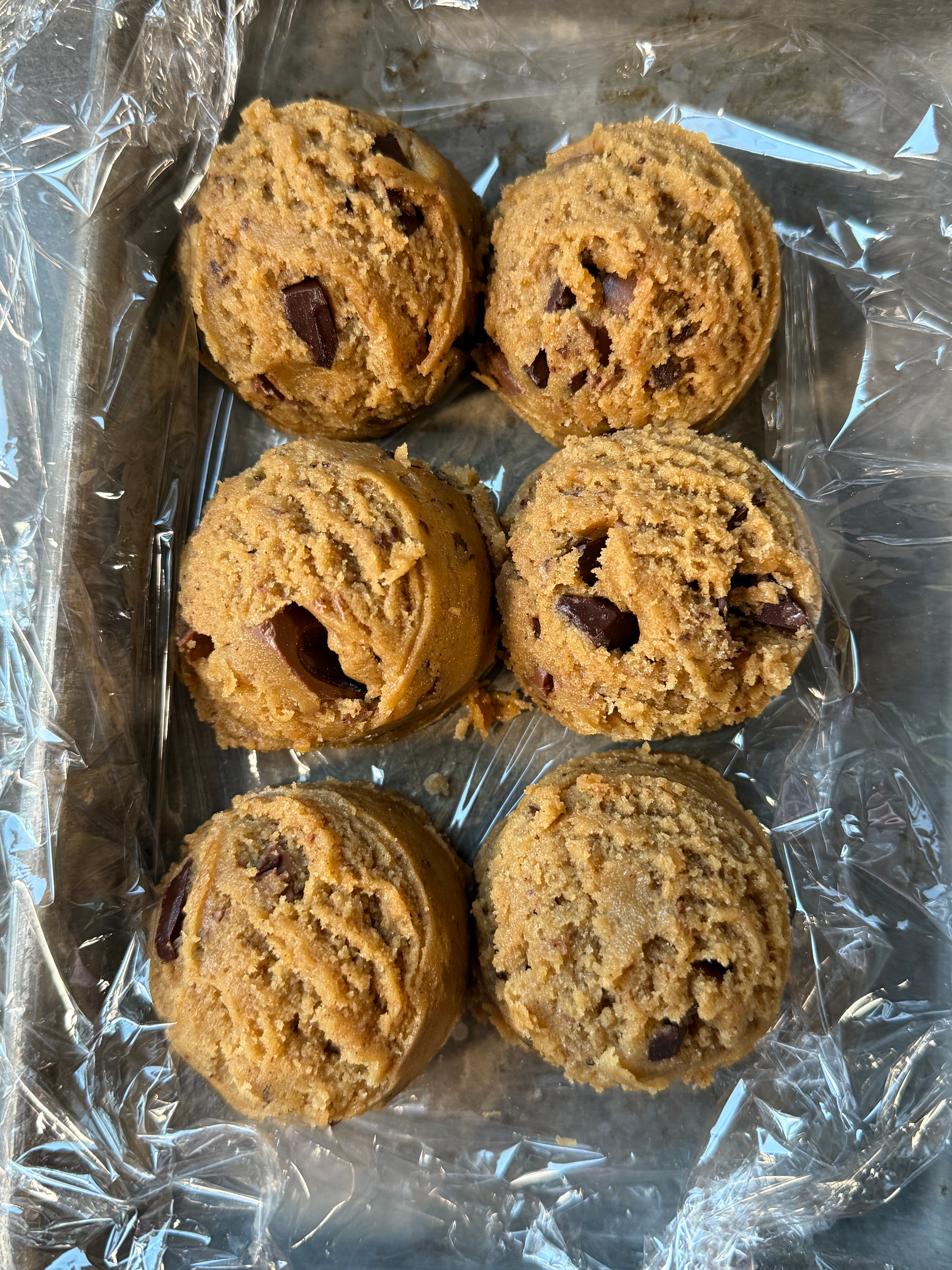 Six scoops of chocolate chip cookie dough on a plastic-lined tray, ready for baking