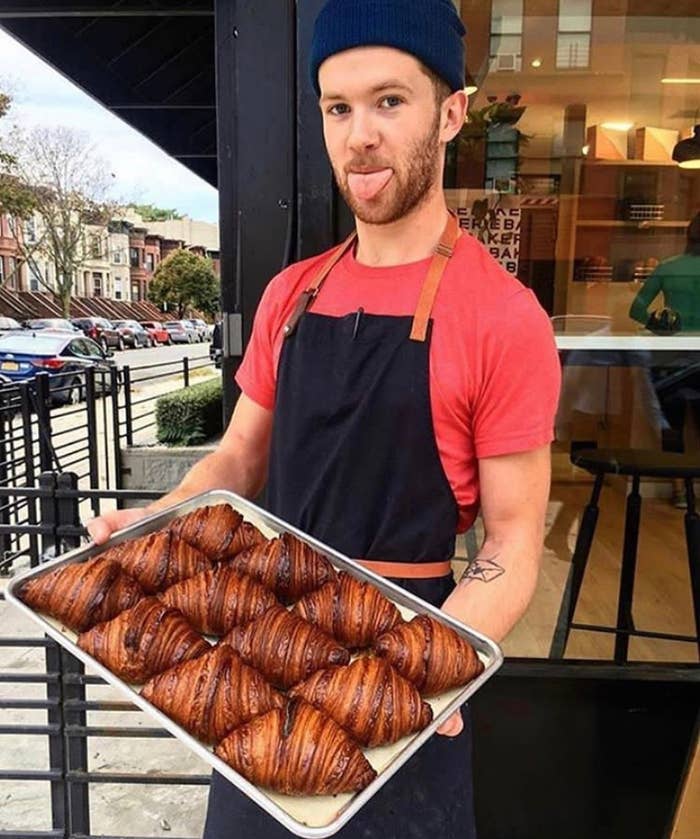 Person in an apron playfully sticks out tongue while holding a tray of croissants outside a bakery