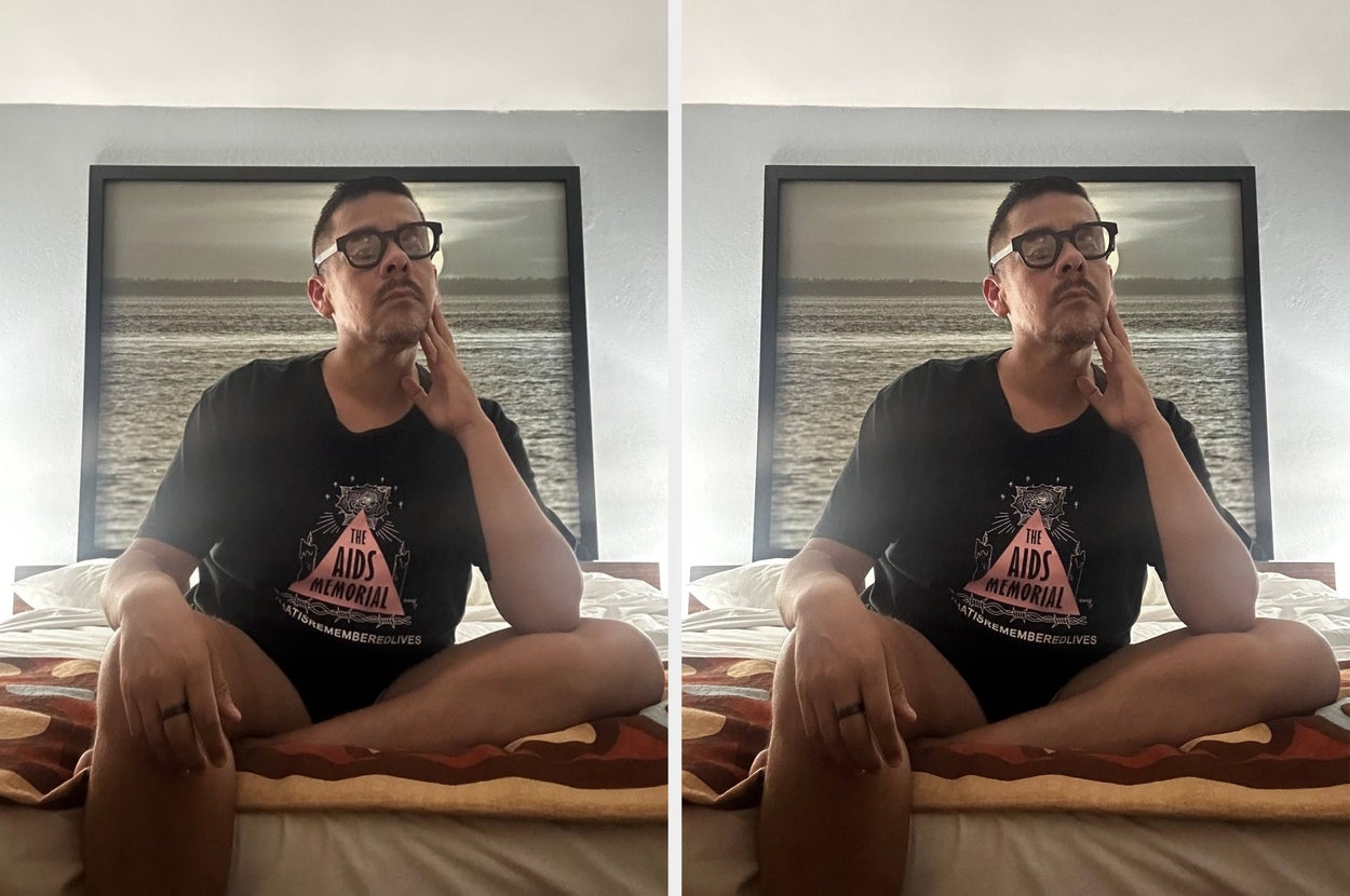 Person in a casual setting, wearing a T-shirt with "The AIDS Memorial" text, seated cross-legged on a bed, reflecting thoughtfully