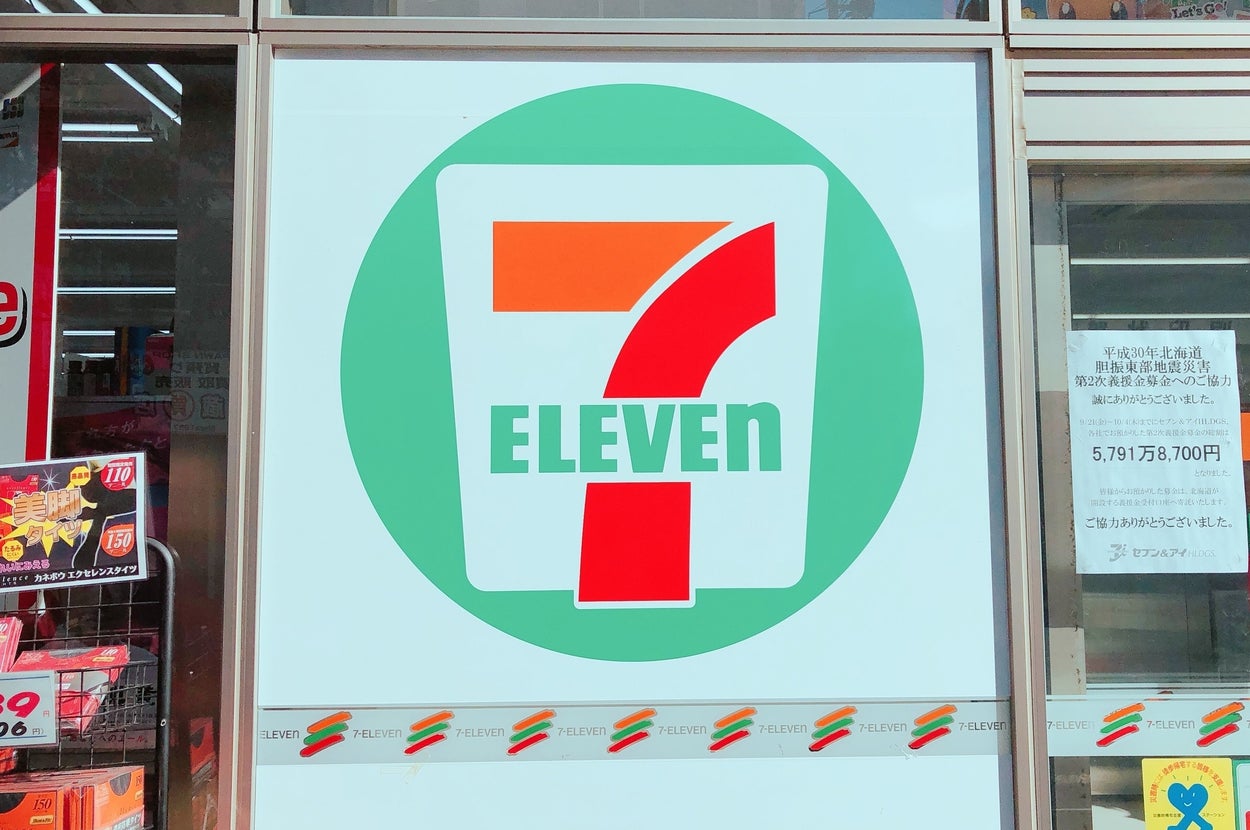 セブンイレブンの店舗外観。大きなロゴが窓に表示されています。商品の広告看板も見えます。