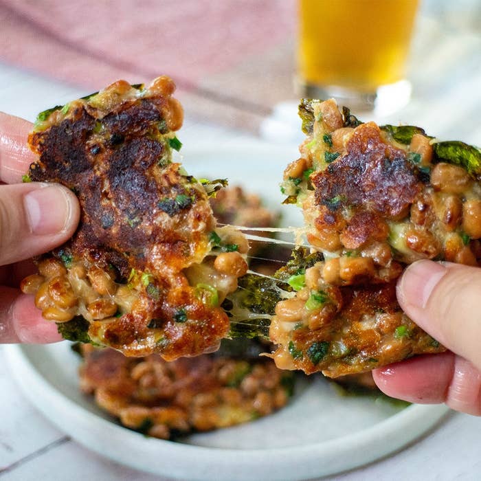手で小さな納豆のお好み焼きを裂く様子。後ろに飲み物が置かれている。