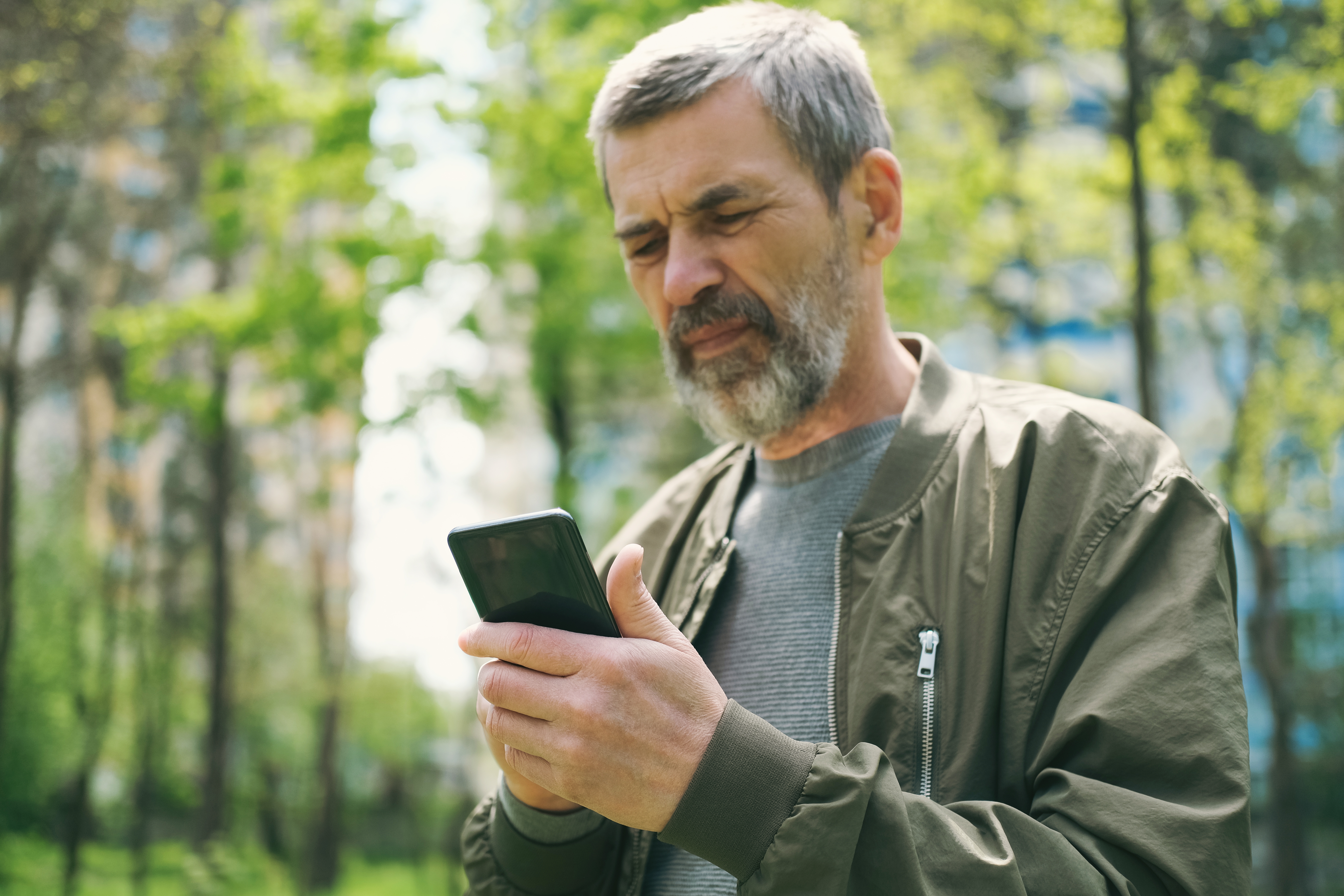 En mann utendørs, holder en smarttelefon og ser på den med et gjennomtenkt uttrykk, iført en uformell jakke og genser