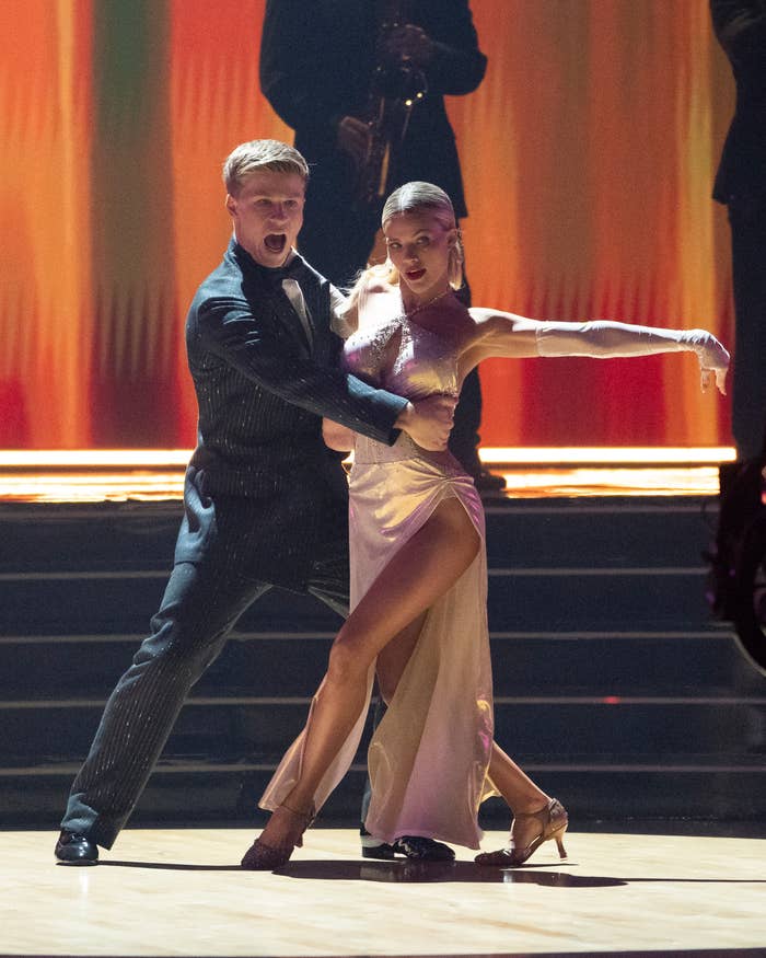 Dancers perform a passionate routine on stage, with one wearing a formal suit and the other in an elegant dress with a high slit