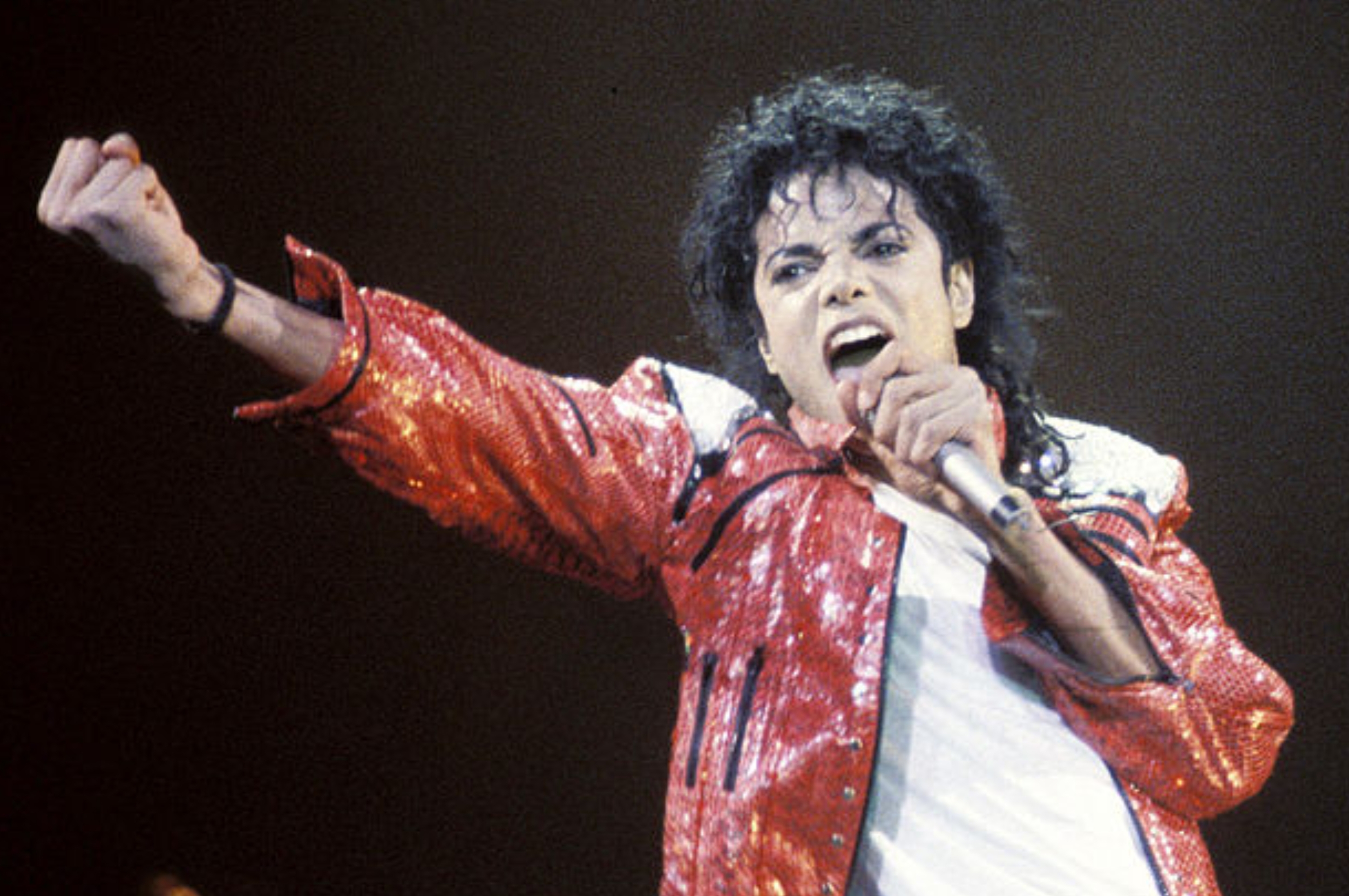 Performer in a sequined jacket energetically sings into a microphone on stage, with an arm outstretched, embodying a dynamic stage presence