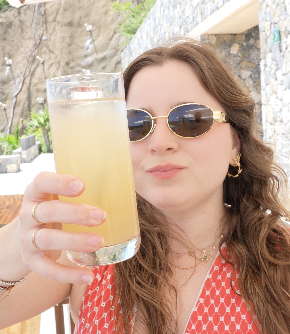 Person outdoors, holding a tall glass with a drink, wearing sunglasses and a patterned top, smiling at the camera